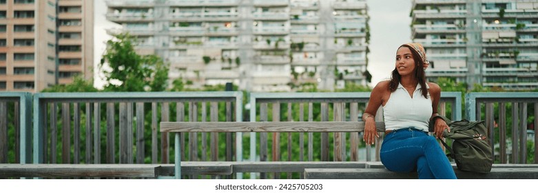 Cute tanned woman with long brown hair wearing white top and yellow bandana seated on bench on cityscape background, Panorama - Powered by Shutterstock