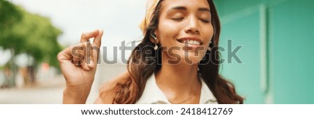 Cute tanned woman with long brown hair wearing white top and yellow bandana listens to music in wireless headphones and dances on blue wall background, Panorama