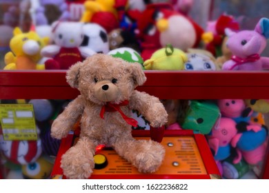 A cute teddy bear with a red scarf sits on a slot machine with his paw on the joystick against the background of soft toys.