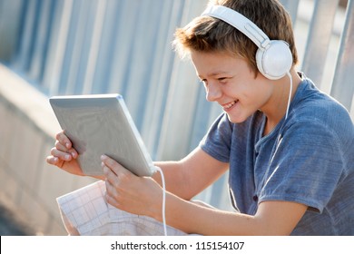 Cute Teen Boy Listening To Music With Headphones And Tablet Outside.