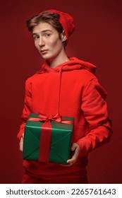 A cute teen boy in a red hoodie and a red hat holds a green gift box and is surprised, making big eyes. Studio portrait on a red background. Christmas and New Year celebration. 