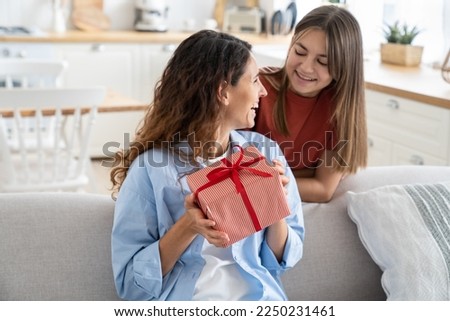 Cute teen girl loving daughter congratulating happy young mother with birthday at home, give her gift box with red ribbon. Child surprising mom on Mothers Day, kid making mum happy. Family holidays 