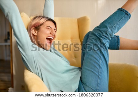 Cute teen girl with nose piercing and pink hair raising hands and feet while sitting comfortably in armchair closing eyes and screaming excitedly, happy with good news, celebrating success