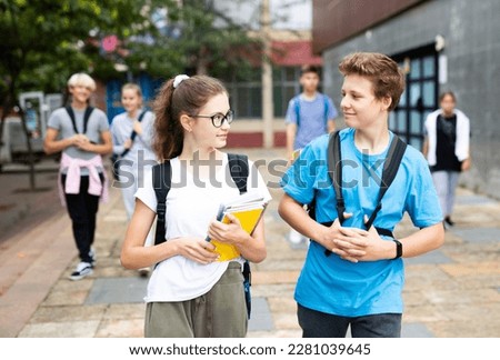 Cute teen girl walking with her classmate outside college building on autumn day, going to lessons.