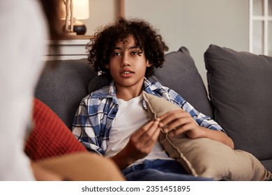 Cute teenage boy sitting on a sofa with pillows and gesturing while talking.