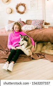 Cute Teenage Girl With Blond Curly Hair At Home, On The Bed In The Loft With A Big Malamute Dog. People And Dogs.
