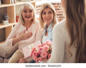 Cute Teenage Girl Is Giving Flowers And A Gift Box To Her Beautiful Granny And Mother While Those Are Sitting On Couch And Smiling