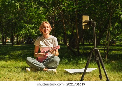 Cute teenage girl playing ukulele in front of the camera. Online music lessons. Relaxation music broadcast on social networks. A girl learns to play the guitar over the Internet while sitting in park - Powered by Shutterstock