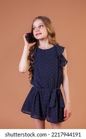 Cute Teenage Girl Princess 10 Year Old With Curly Long Hair Call On Mobile Phone At Beige Background, Talking And Smiling. Fashion Model In Stylish Casual Blue Dress. Fashionable Young Lady Model