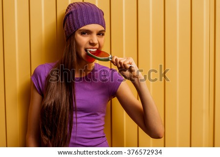 Cute teenage girl in a t-shirt and a cap eating a lollipop, looking in camera and posing, standing on an orange background