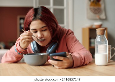 Cute teenager eating cornflakes with milk from bowl and looking at screen of smartphone while sitting by kitchen table and watching video - Powered by Shutterstock