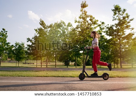 Cute teenager girl riding electric kick scooter in the park at sunset                                       