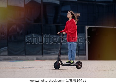 Cute teenager girl riding electric kick scooter in a cityscape                                      