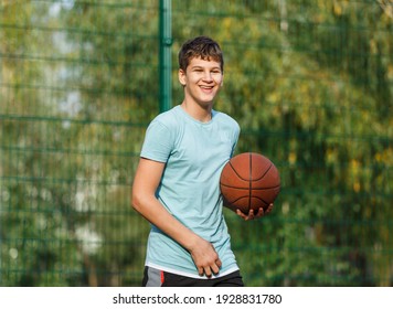 Cute Teenager In Green T-shirt With Orange Basketball Ball Plays Basketball On Street Playground In Summer. Hobby, Active Lifestyle, Sports Activity For Kids.	