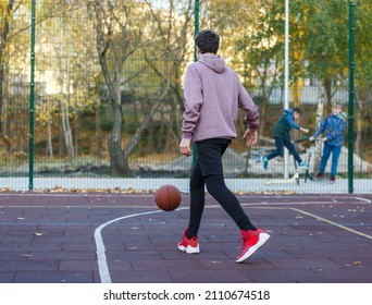 Cute Teenager In A Hoodie Playing Basketball. Young Boy With Ball Learning Dribble And Shooting On The City Court. Hobby For Kids, Active Lifestyle	