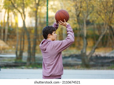 Cute Teenager In A Hoodie Playing Basketball. Young Boy With Ball Learning Dribble And Shooting On The City Court. Hobby For Kids, Active Lifestyle	