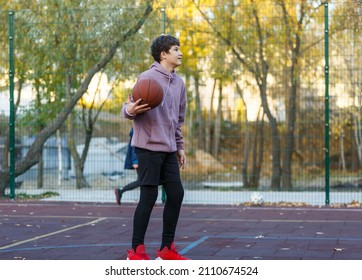 Cute Teenager In A Hoodie Playing Basketball. Young Boy With Ball Learning Dribble And Shooting On The City Court. Hobby For Kids, Active Lifestyle	