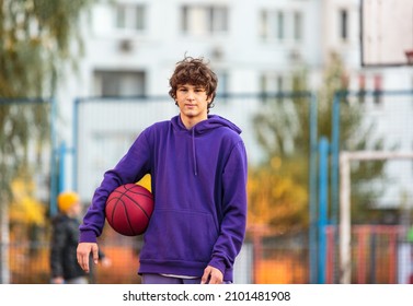 Cute Teenager In Violet Hoodie Playing Basketball. Young Boy With Ball Learning Dribble And Shooting On The City Court. Hobby For Kids, Active Lifestyle