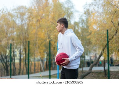 Cute Teenager In White Hoodie Playing Basketball. Young Boy With Red Ball Learning Dribble And Shooting On The City Court. Hobby For Kids, Active Lifestyle	
