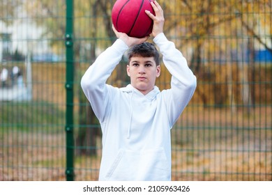 Cute Teenager In White Hoodie Playing Basketball. Young Boy With Red Ball Learning Dribble And Shooting On The City Court. Hobby For Kids, Active Lifestyle	