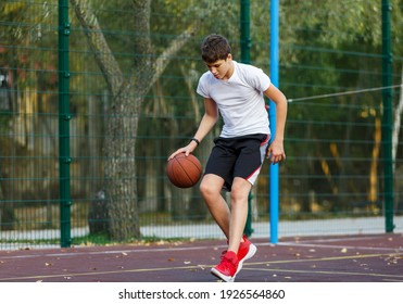  Cute Teenager in white t-shirt with orange basketball ball plays basketball on street playground in summer. Hobby, active lifestyle, sports activity for kids.	