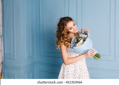 Cute Tender Blond Girl Standing In A Studio With Blue Walls And Holding A Bouquet Of Flowers In Her Hands. She Wears White Dress And Smiles. Flowers Are Wrapped In Blue Paper.