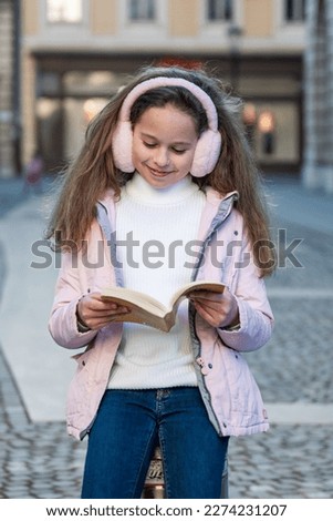 Cute thoughtful girl reading a book outside after school wearing pinl headphones in the city space