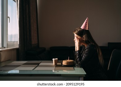 Cute Thoughtful Woman In Party Hat Looking At Festive Cake With Candle. Lonely Woman Freelancer Celebrating Birthday Without Friends. Quarantine And Stay At Home Concept