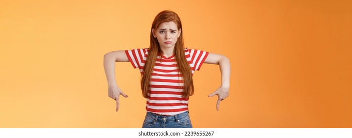 Cute timid hesitant ginger female taking tough decision feel pressured upset, frowning, pulling gloomy unhappy face, pointing down disappointed, uneasy taking decision, orange background. - Shutterstock ID 2239655267