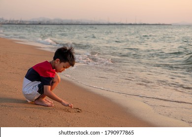 Cute Toddler Asian Boy Sitting Alone On The Beach At Sunset . Child Sitting 
On The Beach Near Sea And Writing On The Sand, Silhouette Of The Boy On The Seaside In Sunset.