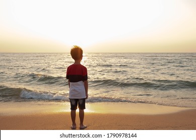 Cute toddler Asian boy standing alone on the beach at sunset. Child standing on the beach near sea and looking on the waves, silhouette of the boy on the seaside in sunset.