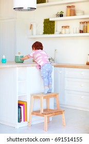 Cute Toddler Baby Climbs On Step Stool, Trying To Reach Things On The High Desk In The Kitchen