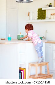 Cute Toddler Baby Climbs On Step Stool, Trying To Reach Things On The High Desk In The Kitchen