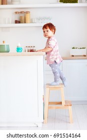 Cute Toddler Baby Climbs On Step Stool, Trying To Reach Things On The High Desk On The Kitchen