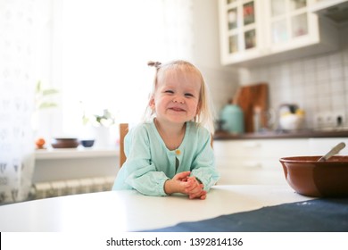 Cute Toddler Blonde Girl Sitting At The Table In The Kitchen, Argues. Light Real Interior