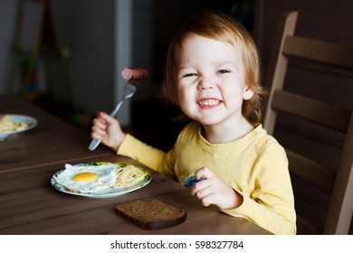 Cute Toddler Boy Eating Sausage And Eggs For Breakfast.