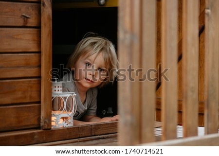 Cute toddler boy, holding lantern, hiding behind wooden door in little playhouse in garden