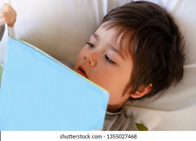 Cute Toddler Boy Laying In Bed Readinga Book Before He Goes To Sleep. Child Reading Story Before Bed, Bed Time Stories Is A Good Habits For Family Spending Time Together.