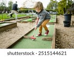 Cute toddler boy playing mini golf having fun on a playground outdoors on warm autumn day. Active leisure for kids in fall.