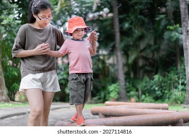 Cute Toddler Boy Walking On Balance Beam With Her Mother's Assistance In A Public Park