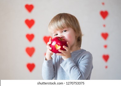 Cute toddler child smiling and eating  red apple in hands on the day of valentine on the background of hearts