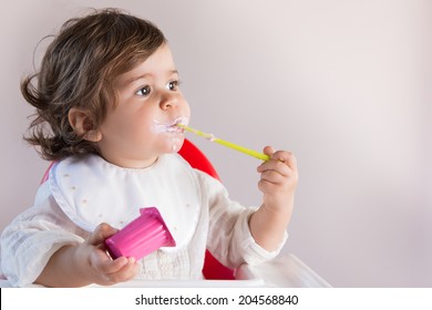 Cute Toddler Eating Yogurt While Looking At. Her Pretty Face Has Yogurt Around Her Mouth.