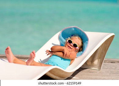 cute toddler girl in bikini, hat and sunglasses lyiing on a lounge and relaxing in tropic ocean background