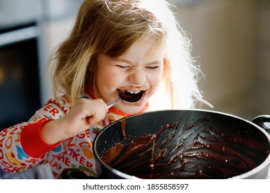 Cute Toddler Girl Eating Chocolate Dough Rests With Spoon And Fingers From Pot. Happy Child Licking Sweet Dough For Muffins Or Cake, Helping In Home Kitchen, Indoors.