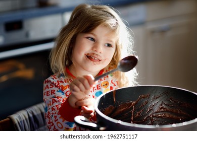 Cute Toddler Girl Eating Chocolate Dough Rests With Spoon And Fingers From Pot. Happy Child Licking Sweet Dough For Muffins Or Cake, Helping In Home Kitchen, Indoors