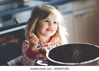 Cute Toddler Girl Eating Chocolate Dough Rests With Spoon And Fingers From Pot. Happy Child Licking Sweet Dough For Muffins Or Cake, Helping In Home Kitchen, Indoors.