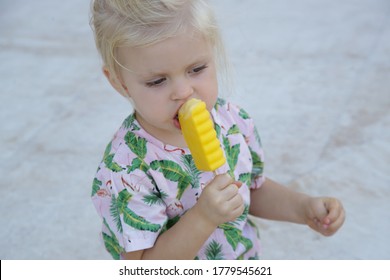 Cute Toddler Girl Eating Yellow Popsicle. Candid Outdoor Portrait Of Child With Ice Cream.