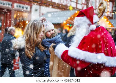 Cute Toddler Girl With Mother On Christmas Market. Funny Happy Kid Taking Gift From Bag Of Santa Claus. Holidays, Christmas, Childhood And People Concept. Happy Family During Snowfall On Winter Day.