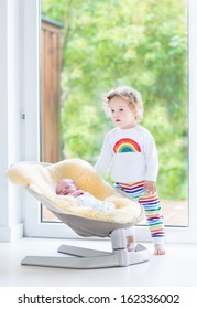 Cute toddler girl playing with her newborn baby brother relaxing in a swing next to a big window and door to the garden