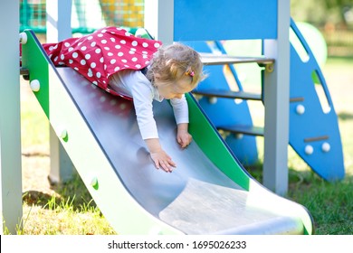 Cute Toddler Girl Playing On Slide On Outdoor Playground. Beautiful Baby In Red Gum Trousers Having Fun On Sunny Warm Summer Day. Child Sliding Down